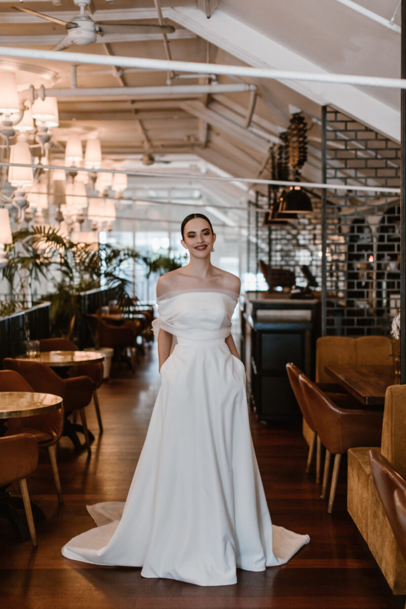 This classic A-line wedding gown flows over the body, with wide off the shoulder detailing & small hidden pockets this gown if made for the timeless bride.