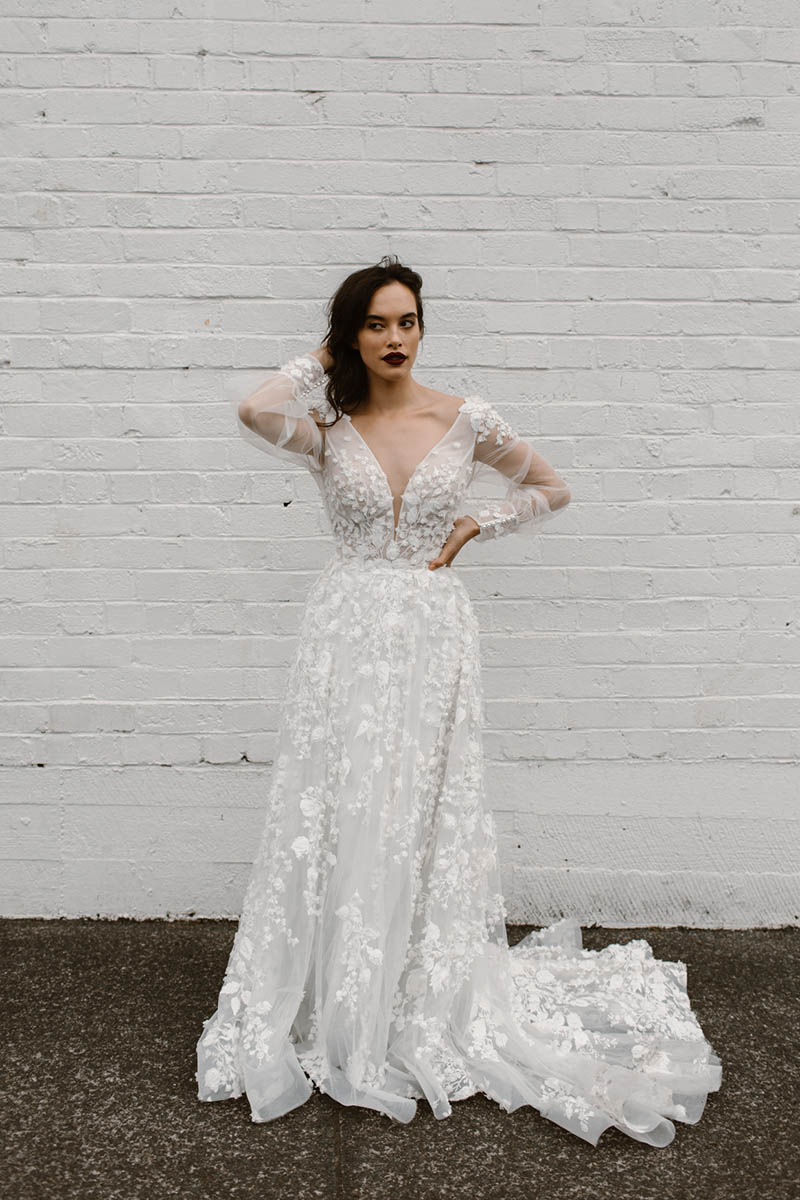 Queeny is a princess wedding dress like no other. Long lace sleeves, 3D flowers, plunging neckline, soft flowing skirt & graceful train. She is breathtaking